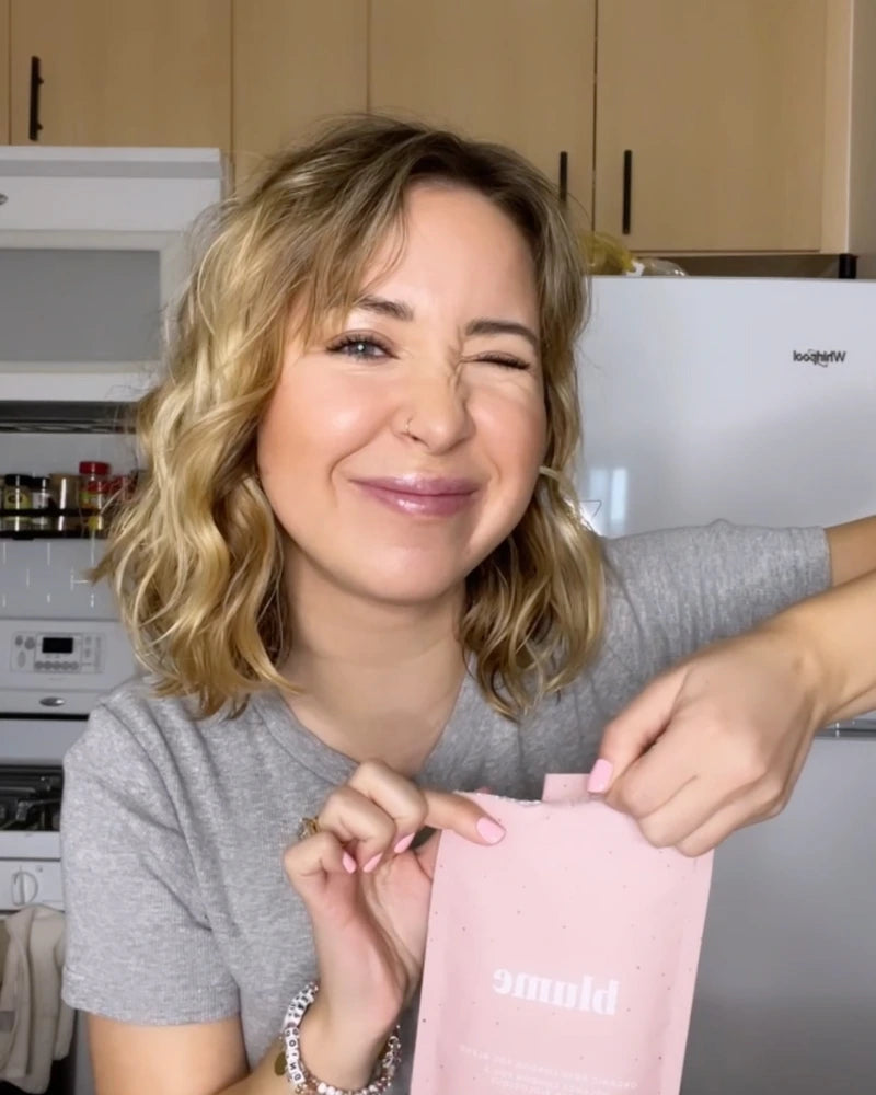 A woman with wavy blonde hair winks at the camera while holding a pink package labeled bloom. She stands in a kitchen with wooden cabinets, a stove, and spices in the background. Shes wearing a gray shirt and bracelet.